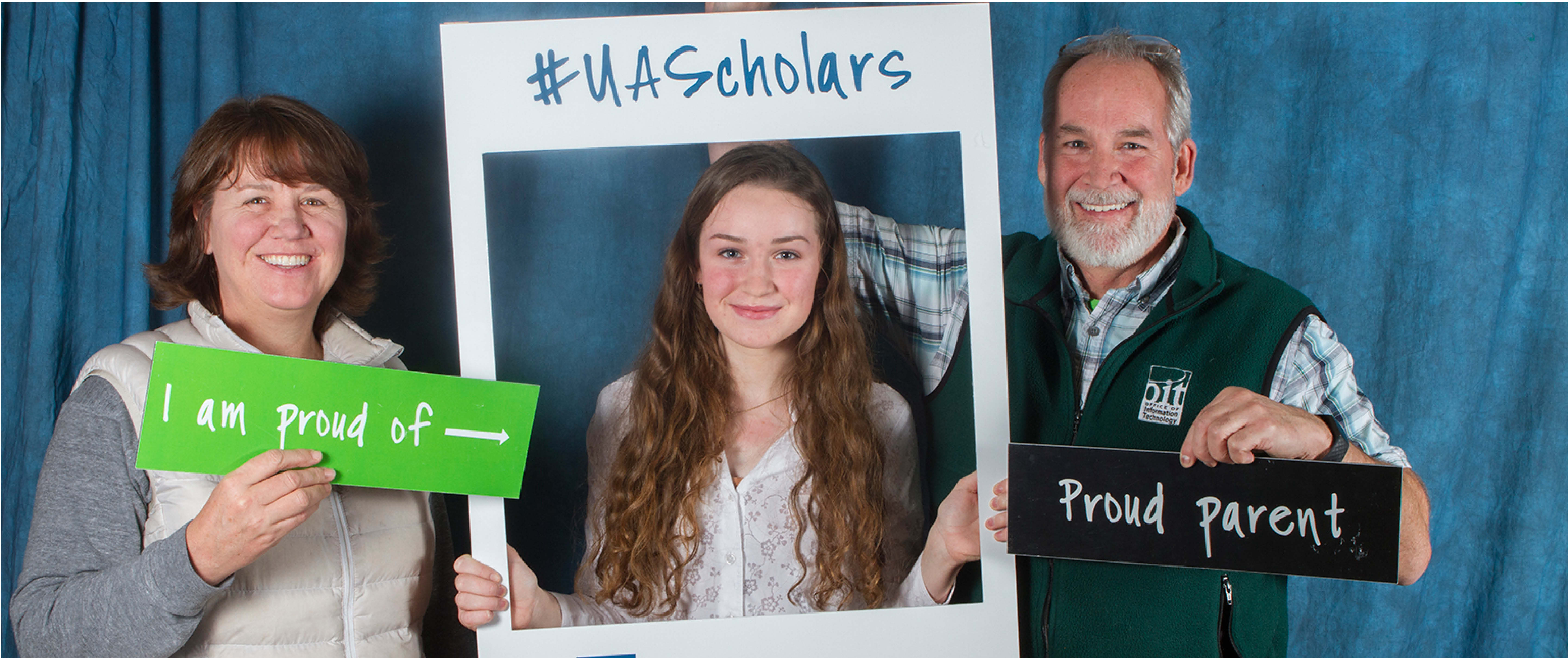 UA Scholar surrounded by mom and dad holding 'proud parent' signs