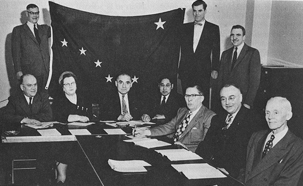 Alaska Statehood Committee: Standing with flag (from left to right): Bob Bartlett, Robert Atwood, Barrie White (non-member). Seated (from left to right): W.L. Baker, Mildred Hermann, Frank Peratrovich, Percy Ipalook, Warren Taylor, Victor Rivers, Andrew Nerland. Not Pictured: Stanley McCutcheon, Lee Bettinger